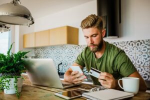 Man sitting in front of laptop holding credit card while using smart phone while working from home izusek eVPBex